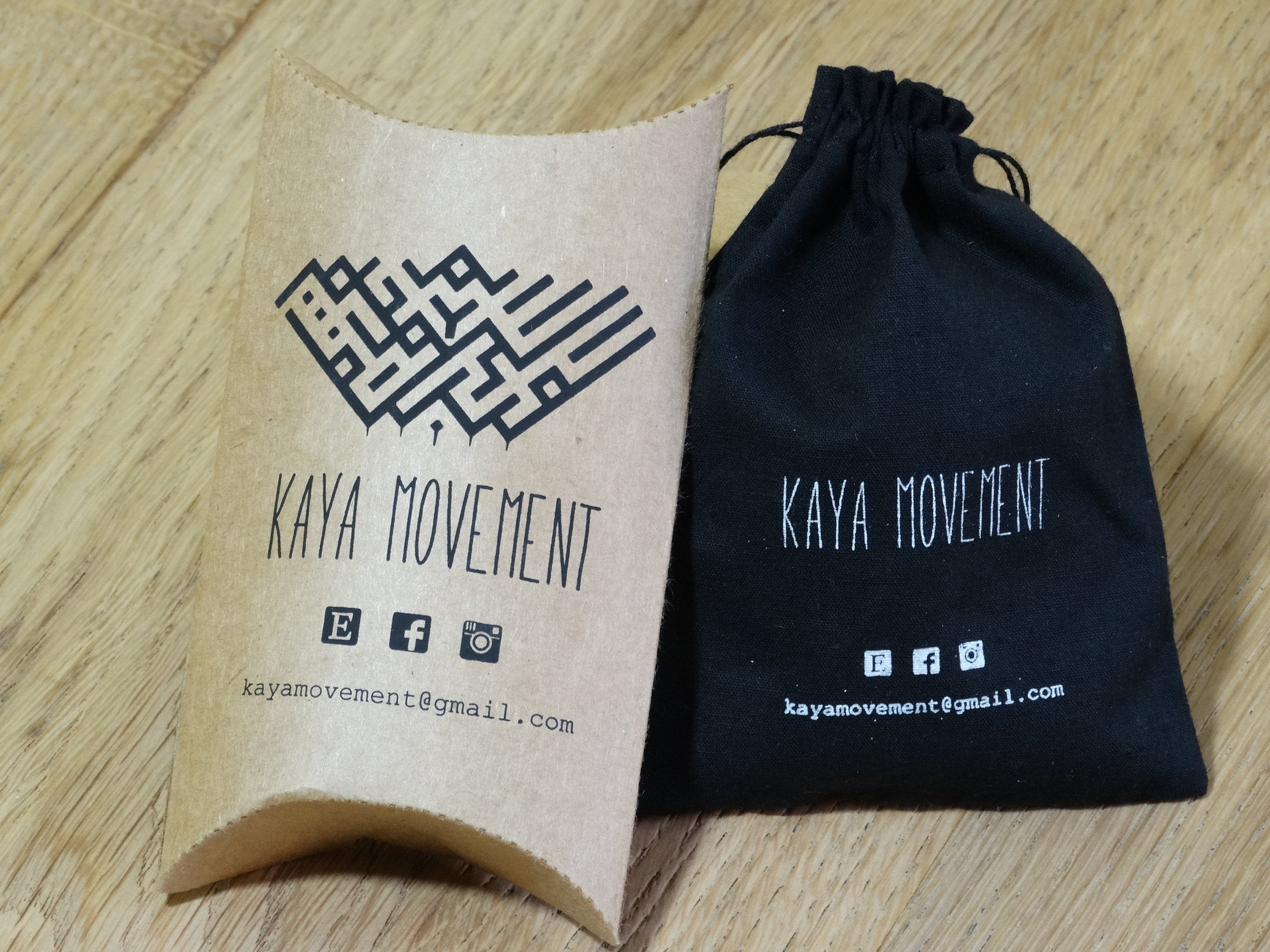 A small, brown, rectangular pillow box and a black drawstring pouch, both labeled "Kaya Movement." The packaging features the website kayamovement@gmail.com and social media icons for Facebook, Instagram, and Twitter. They are placed on a wooden surface alongside the HAMSA HAND Belly Piercing Ring in Gold | 14 gauge surgical steel from Kaya Movement.
